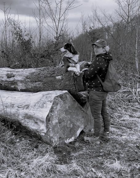 Frau mit Hund auf einem Baumstamm in einer herbstlichen Landschaft.