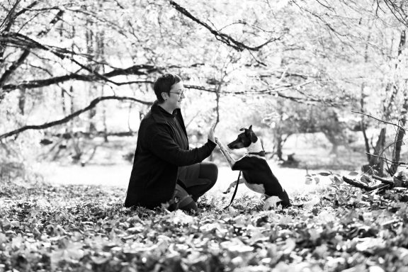 Ein Mann kniet neben einem Hund in einer herbstlichen Landschaft mit Laub.